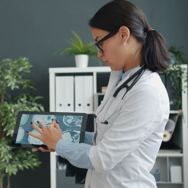 A physician in a white coat points to MRI images on a tablet while discussing them with a patient standing opposite her in a clinic office.