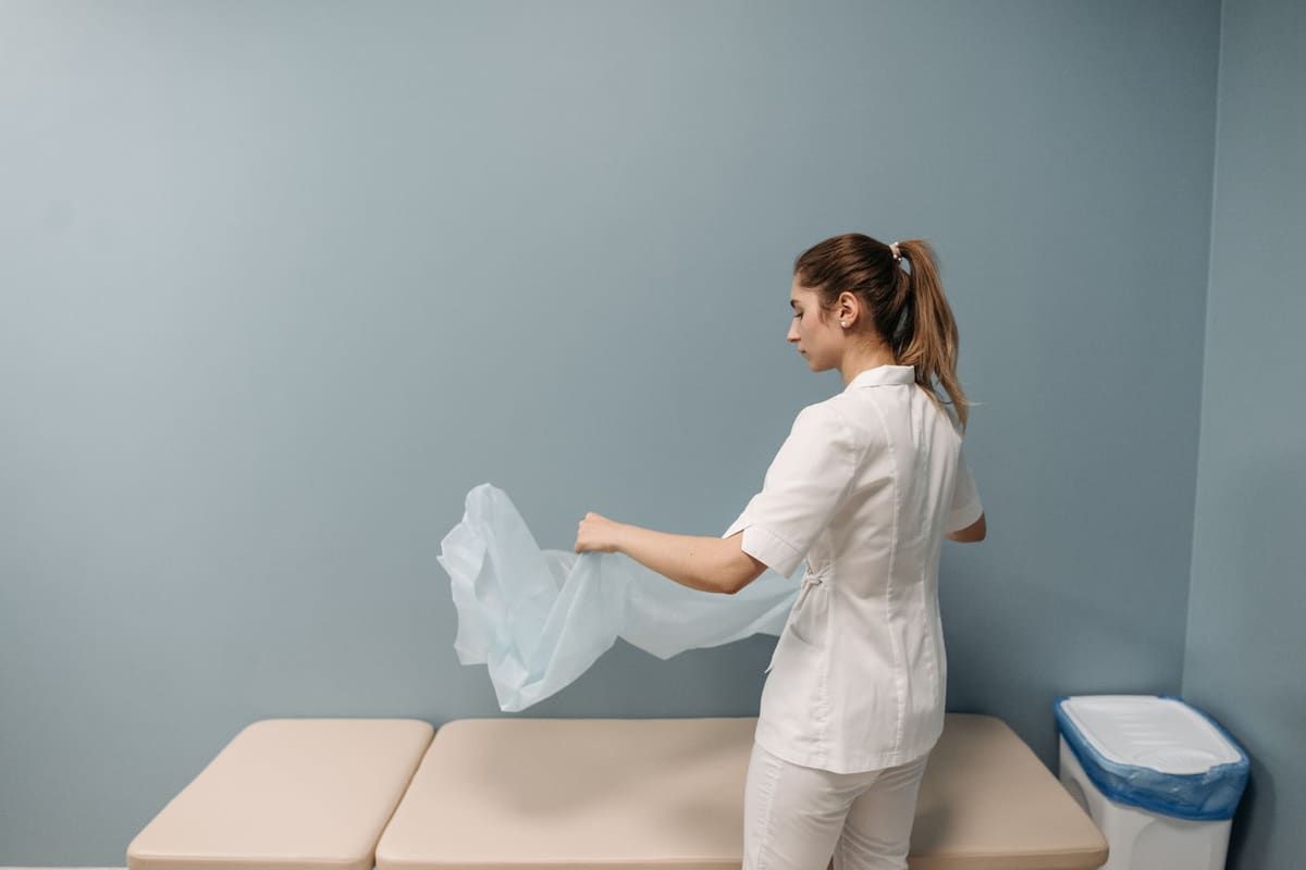 A clinician in white scrubs lays a fresh drape over a treatment table in a quiet outpatient procedure room.