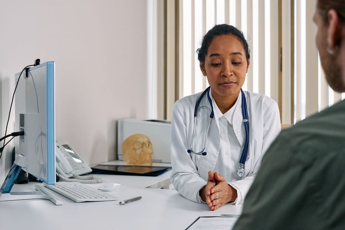 A physician sits across a desk from a patient in a bright medical office, listening during a consultation.
