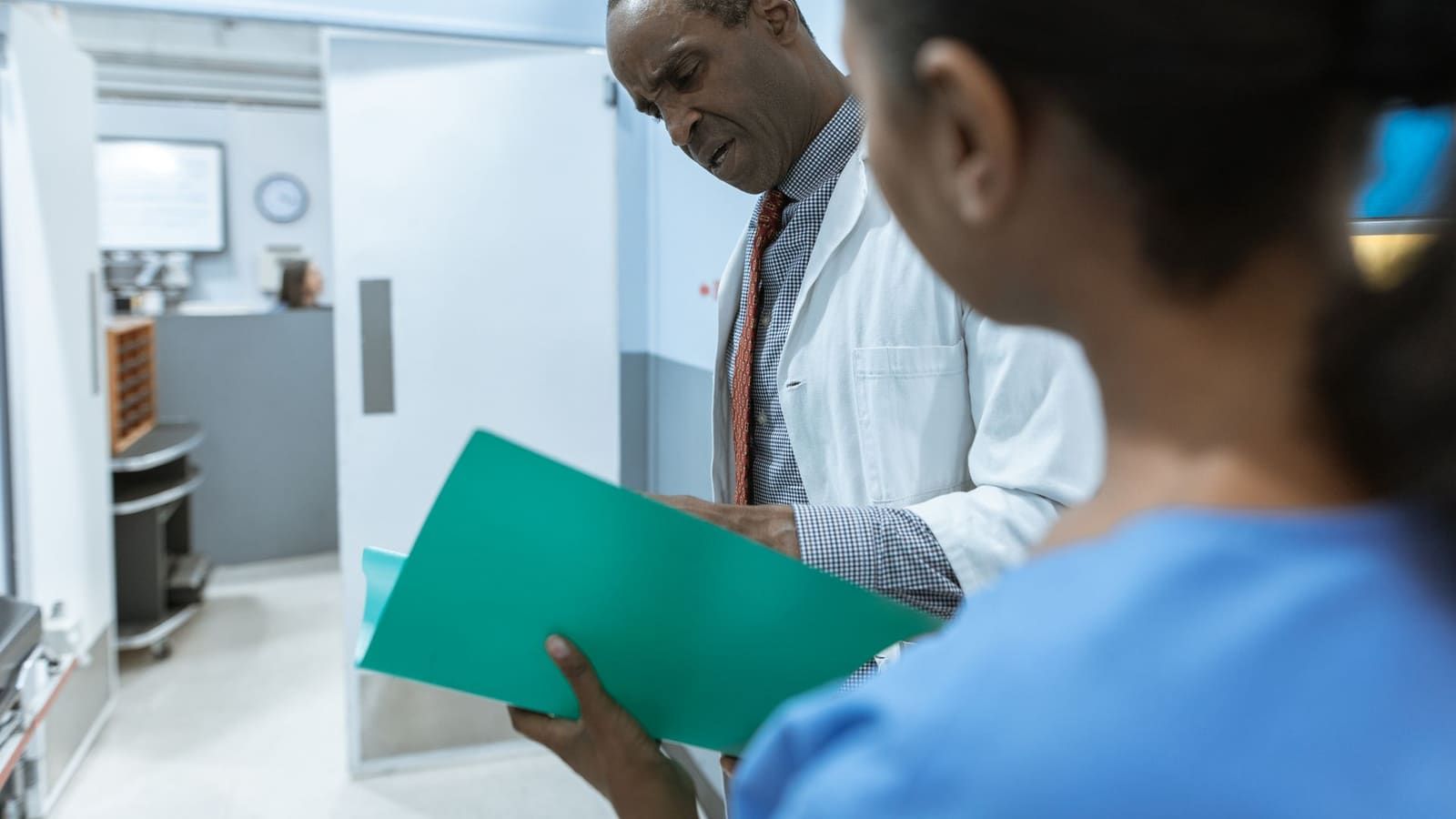 Two clinicians in a bright clinic corridor review a patient chart together in a green folder.