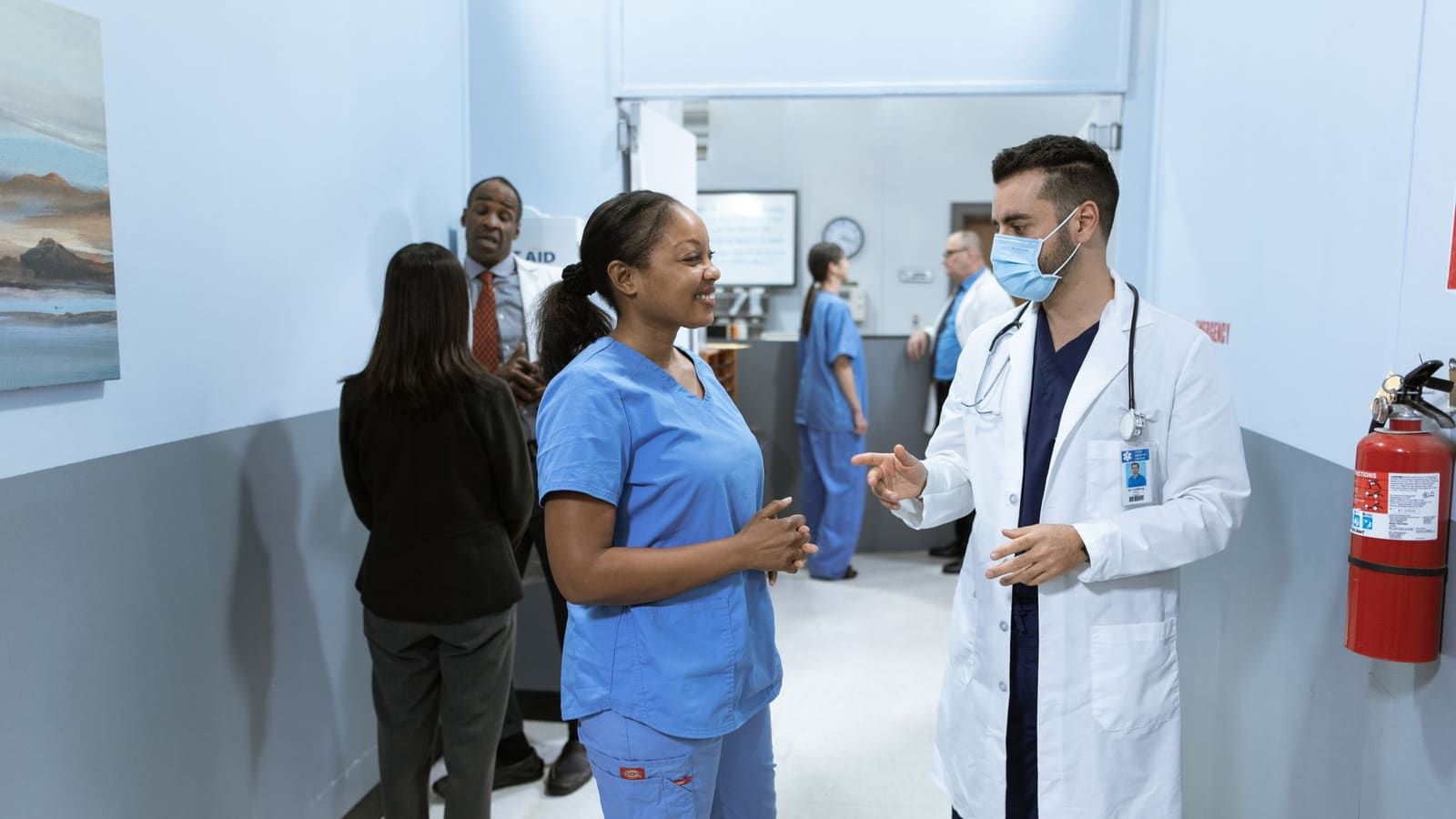 A physician in a white coat and surgical mask talks with a smiling nurse in blue scrubs in a clinic hallway, while other doctors, nurses and staff talk in the background.