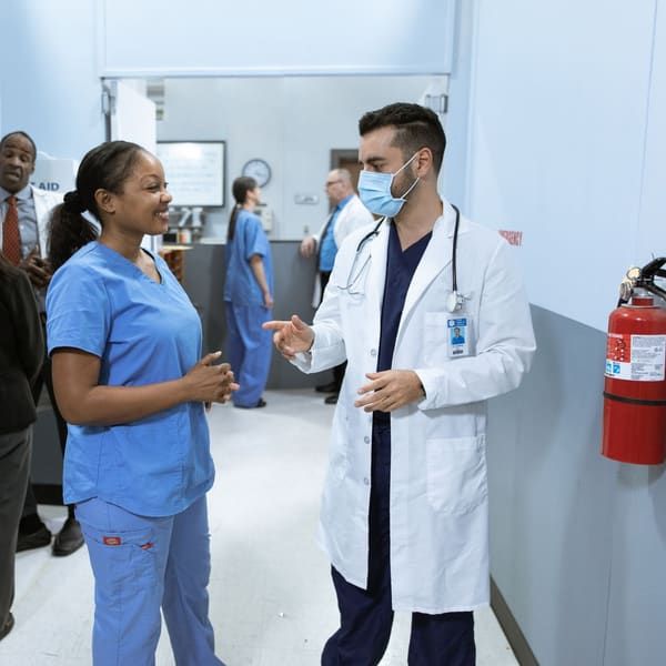 A physician in a white coat and surgical mask talks with a smiling nurse in blue scrubs in a clinic hallway, while other doctors, nurses and staff talk in the background.