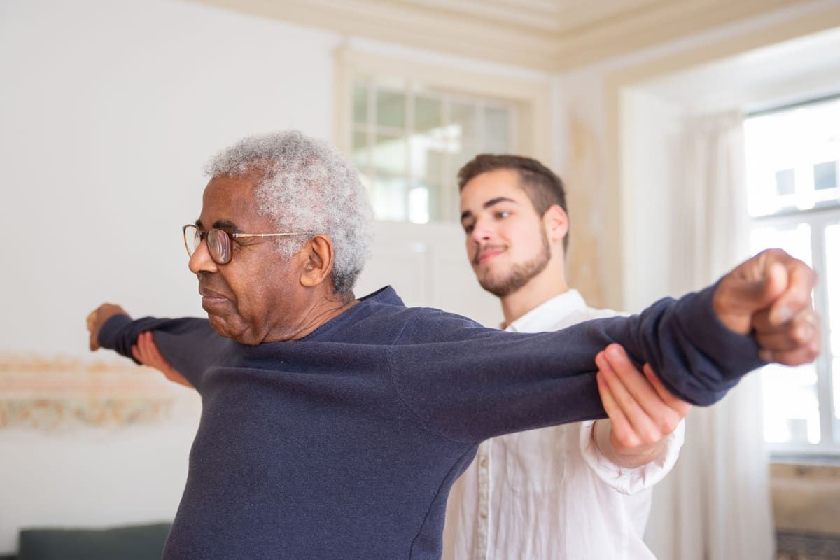 A young man in a white shirt stands behind an older man in glasses and a navy sweater, supporting his outstretched arms as he holds a stretch in a bright room.