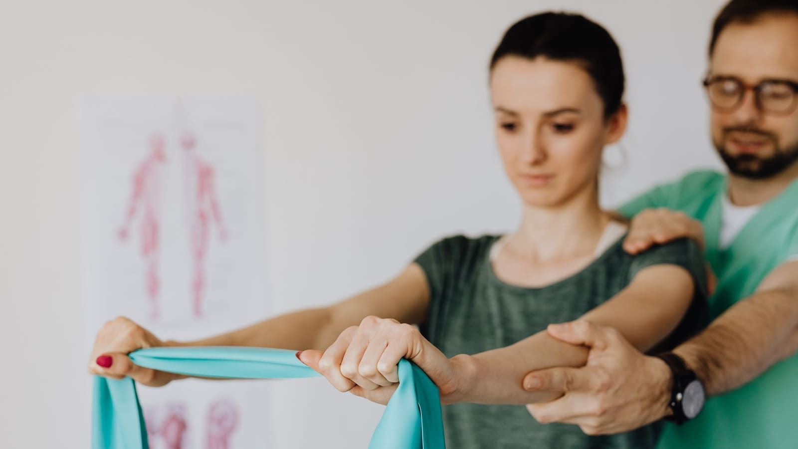 A clinician in green scrubs steadies a patient by the shoulder and forearm as she pulls a teal resistance band with both arms, an anatomy chart on the wall behind them.