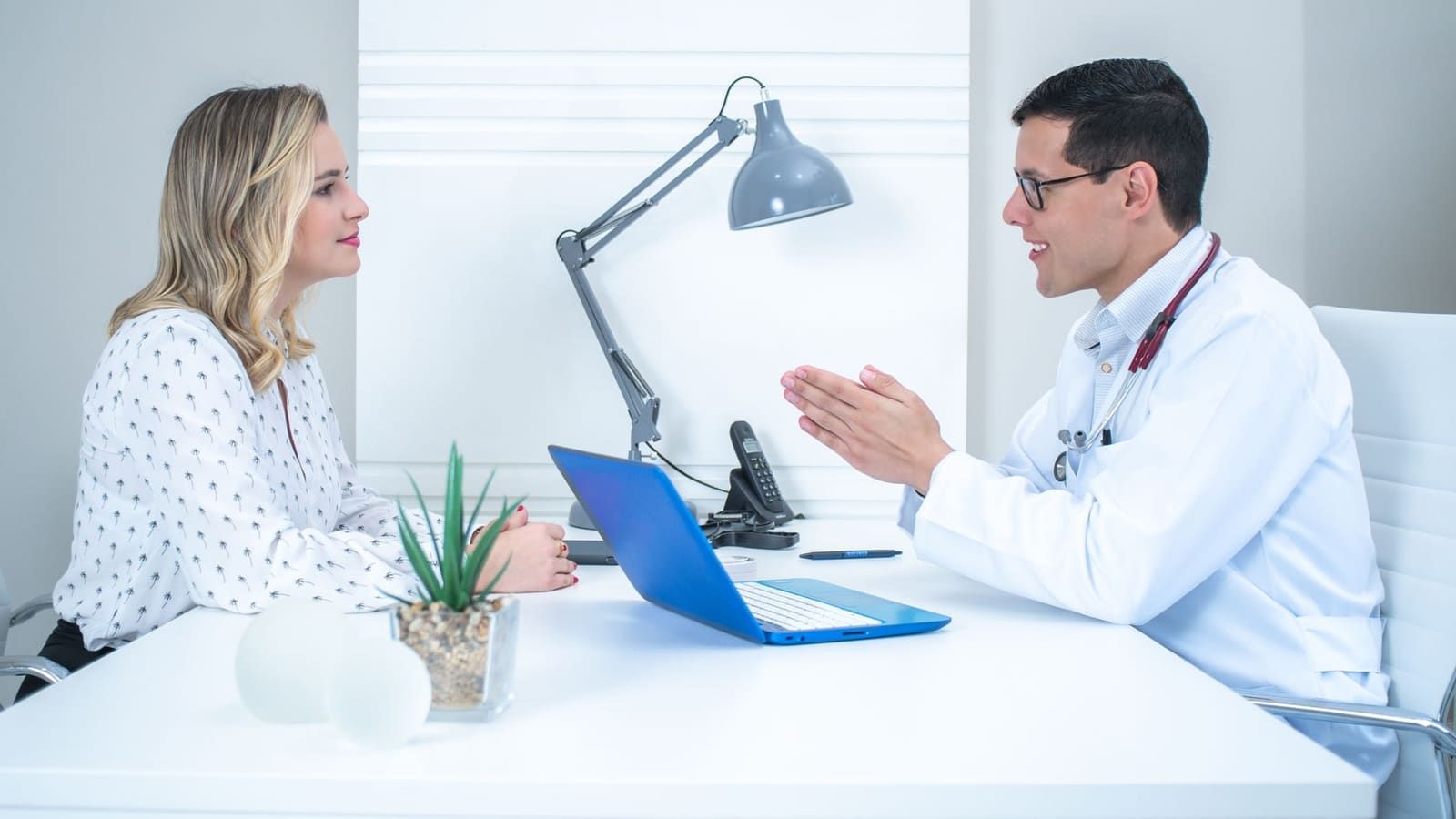 A physician in a white coat gestures as he explains something across a desk to a woman in a patterned blouse, who listens closely, with a laptop and desk lamp between them.