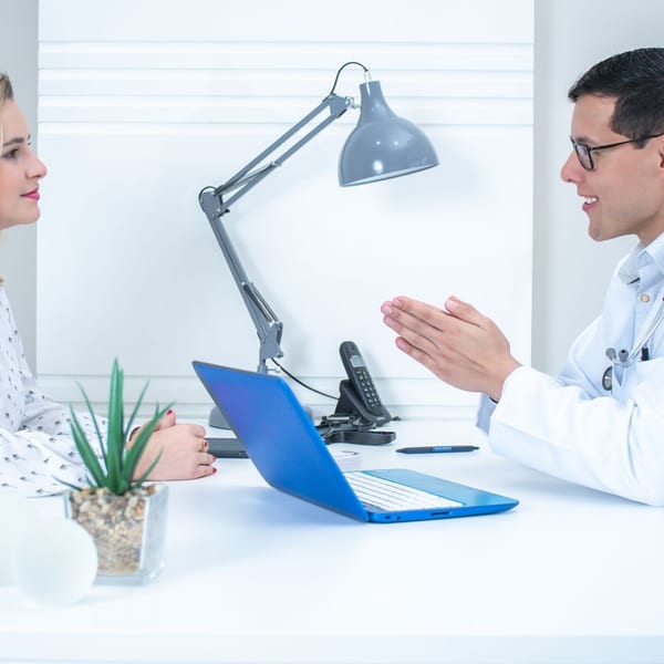 A physician in a white coat gestures as he explains something across a desk to a woman in a patterned blouse, who listens closely, with a laptop and desk lamp between them.