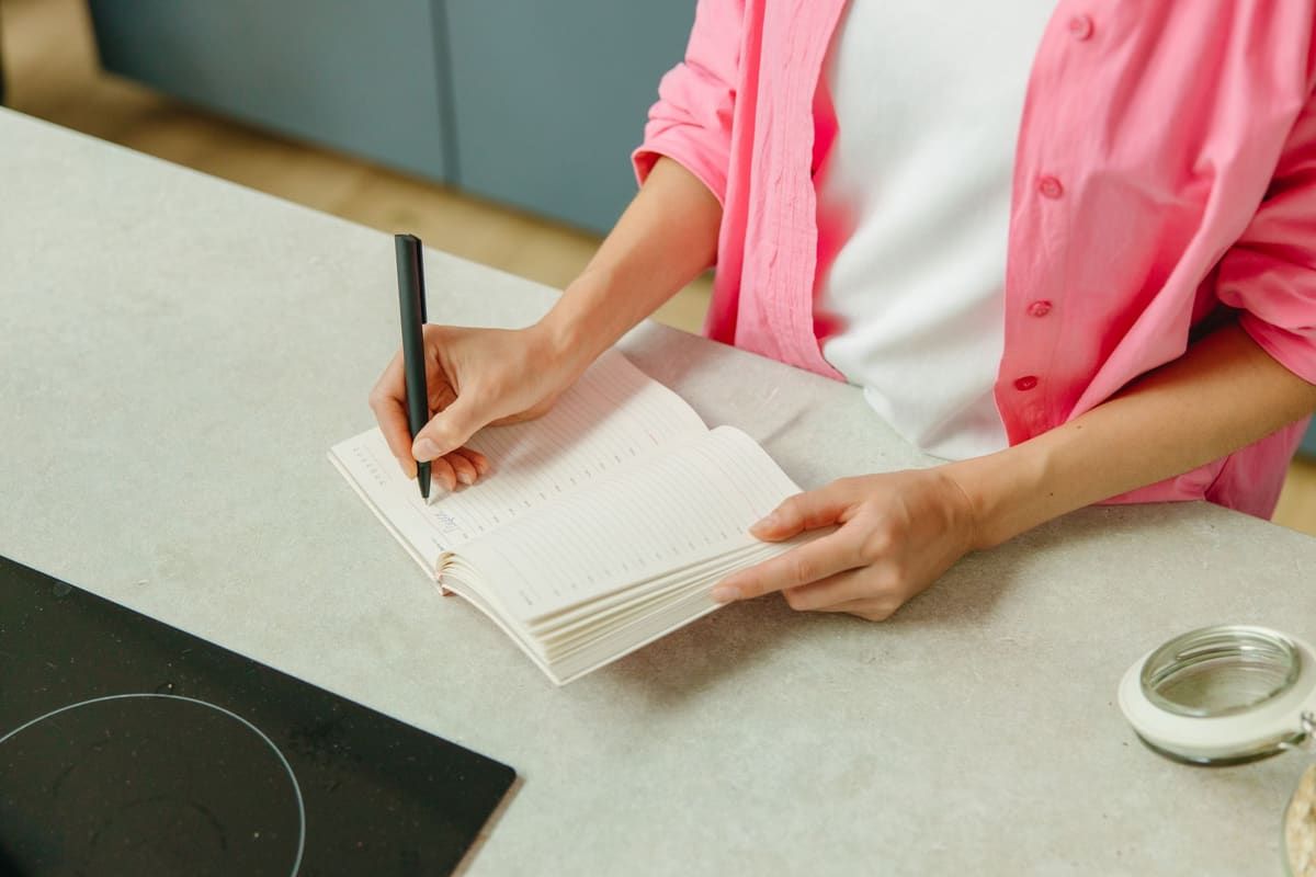 A woman in a pink shirt stands at a kitchen counter writing in an open notebook with a black pen.