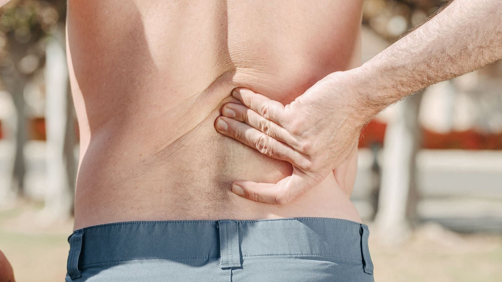 A shirtless man stands outdoors with his back to the camera, pressing his fingers into one side of his lower back just above the waistband of his gray trousers.