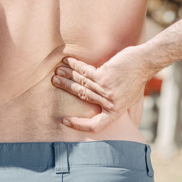 A shirtless man stands outdoors with his back to the camera, pressing his fingers into one side of his lower back just above the waistband of his gray trousers.