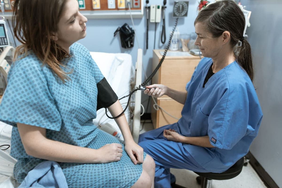A nurse in blue scrubs sits beside a young woman in a patterned hospital gown and takes her blood pressure with an arm cuff in a clinic room.