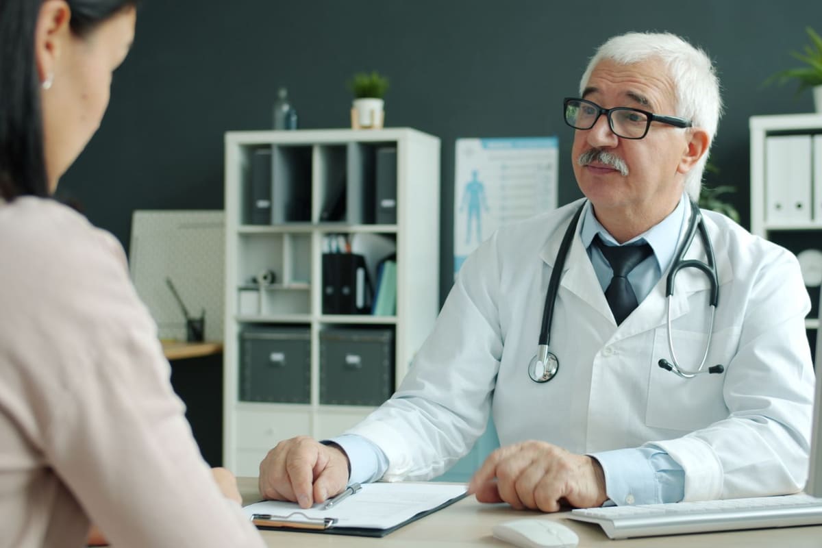 A gray-haired physician in a white coat sits at his desk and listens to a patient seated across from him.