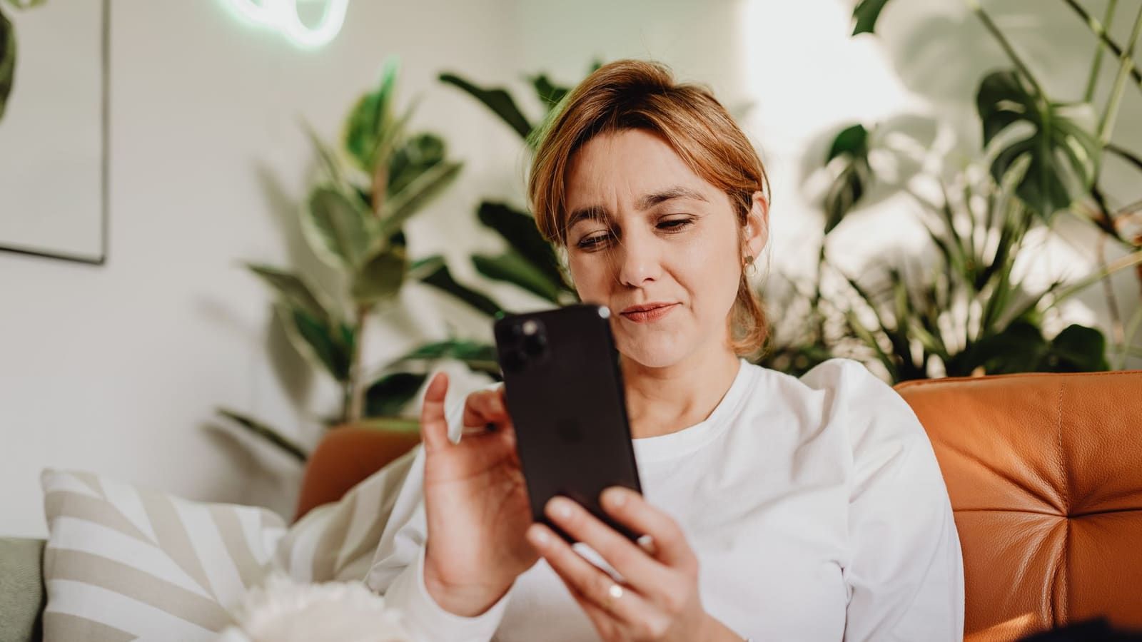 A woman sitting on a leather sofa at home squints skeptically at her smartphone as she reads.