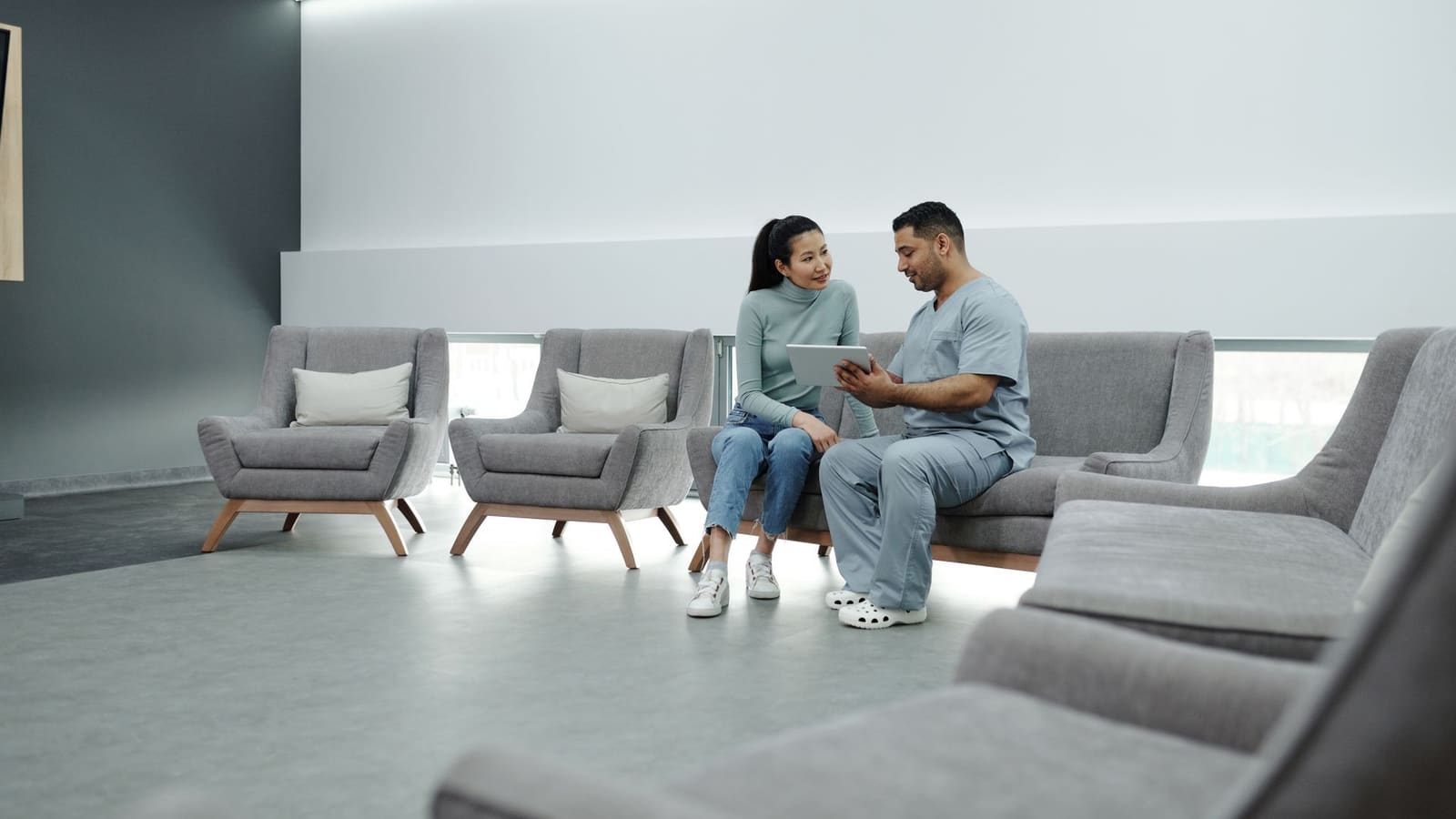 A clinician in scrubs sits with a patient in a bright clinic waiting area, reviewing information on a tablet together.